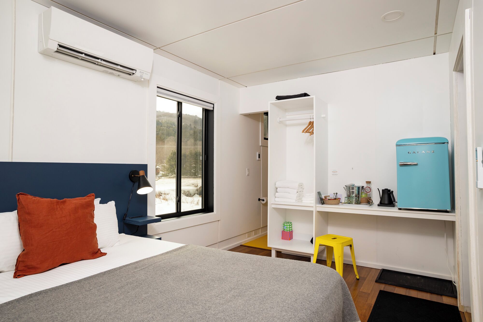 A bedroom with a bed, a window, a wardrobe, a small table, and a blue refrigerator.