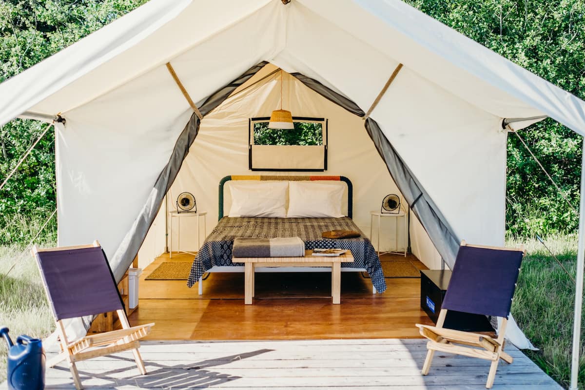 A tent with a bed, two chairs, a small table, and a hanging light fixture.