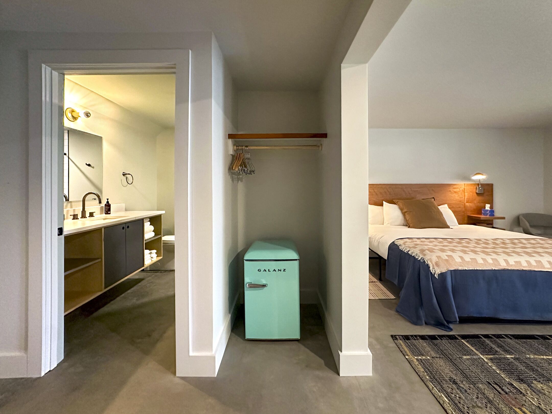 A hotel room with a bed, bedside table, lamp, and a small teal minibar.