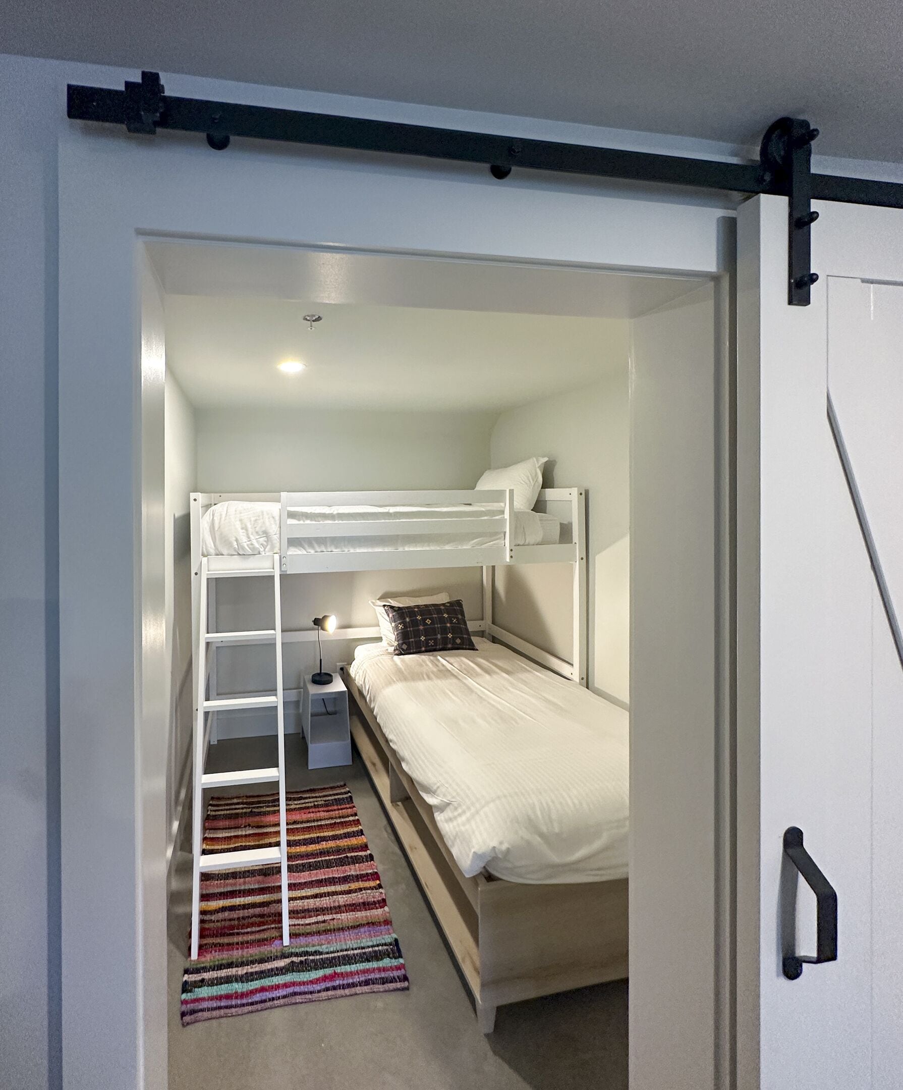 A small, well-lit bedroom with a bunk bed, a bedside table with a lamp, and a colorful striped rug.