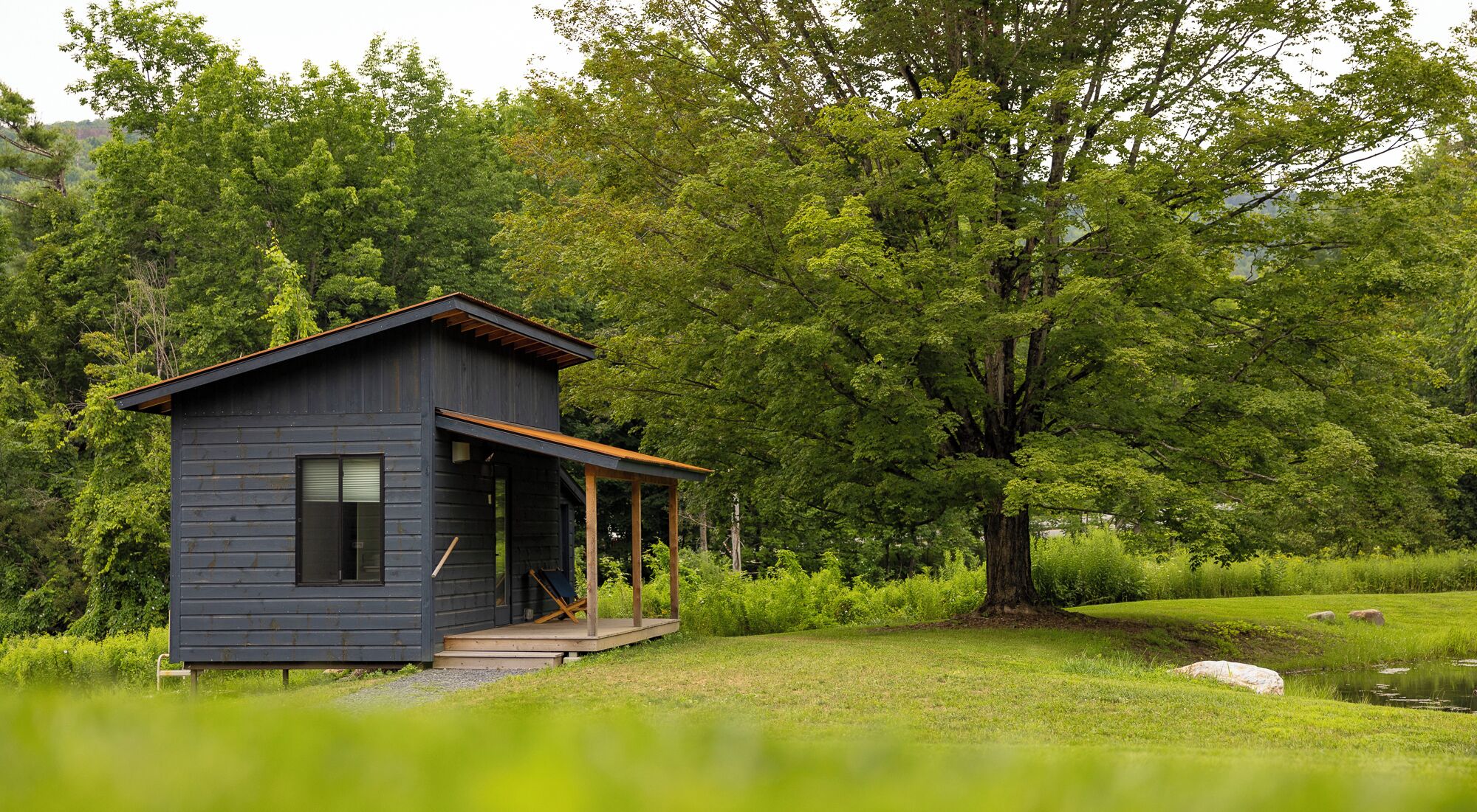 A small, dark wooden cabin with a porch, surrounded by lush greenery and trees.