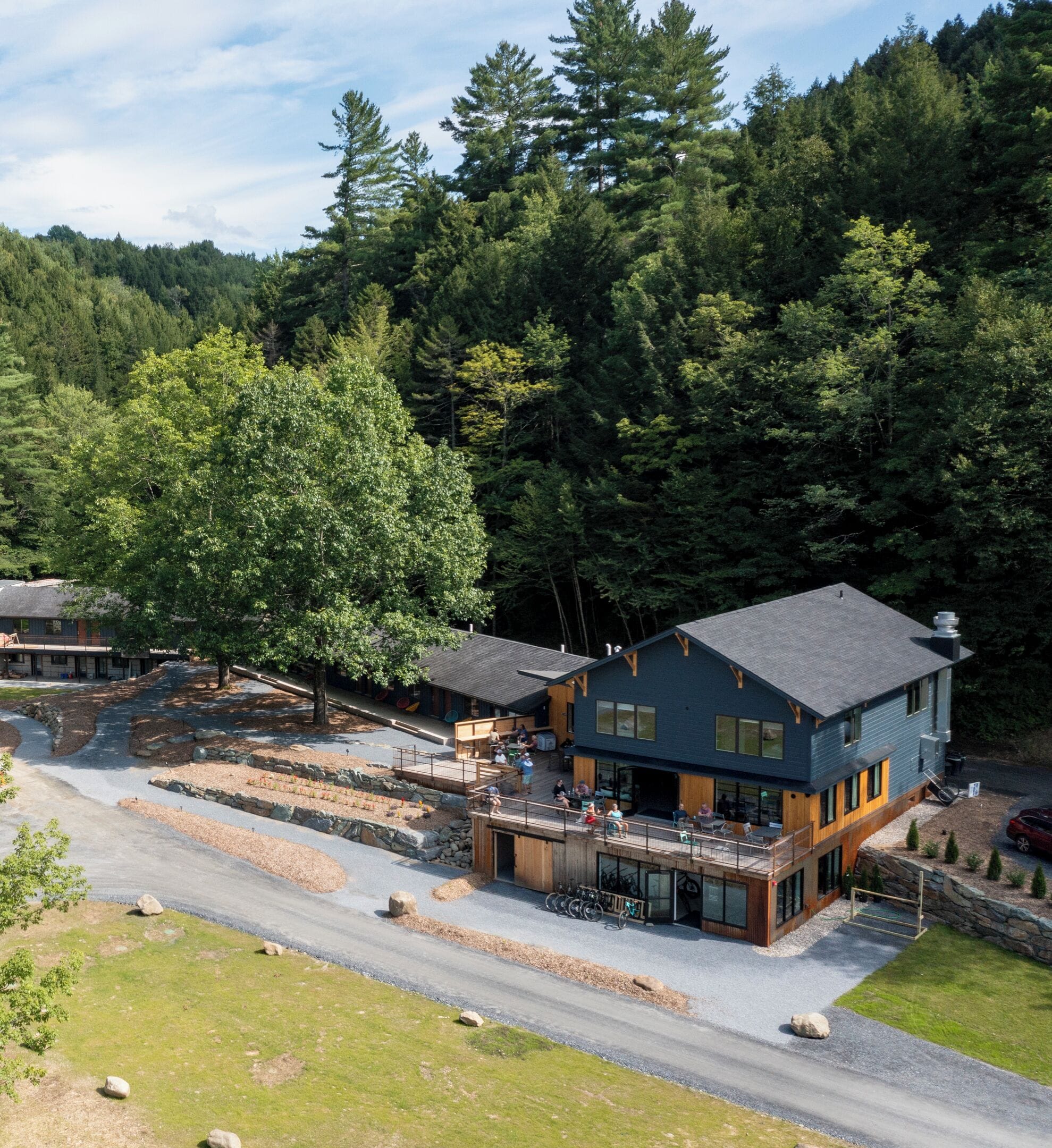 A modern two-story building with a deck and outdoor seating area, surrounded by dense forest.