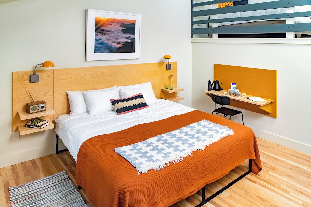 A modern hotel room with a wooden headboard, a bed with white bedding and an orange throw, a desk with a chair, a small shelf with books, a framed picture on the wall, and a bunk bed in the background.
