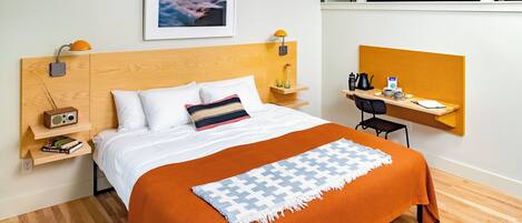 Una habitación de hotel moderna con cabecera de madera, una cama con ropa blanca y una manta naranja, un escritorio con silla, un estante con libros, un cuadro en la pared y una cama litera al fondo.