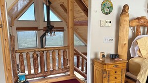 A room with a sloped ceiling, wooden beams, a red carpet, a wooden chest of drawers, a bed, and a view through a window.