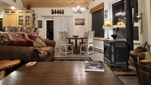 A cozy living room with a wooden ceiling, a fireplace, a dining area with a table and chairs, and a kitchen with wooden cabinets.