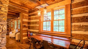 A log cabin interior with a wooden dining table, chairs, and a large window with a view of the forest. - Charming Cabin Near Blue Ridge Mountains (Blowing Rock)