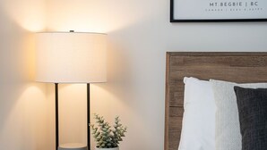 Habitación de hotel ordenada con una cama, una lámpara de noche, una pequeña planta en maceta y un cuadro en la pared.