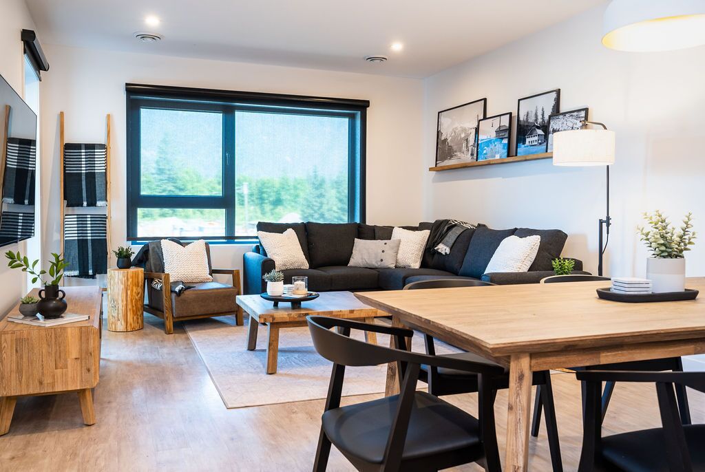 A modern living room with a sofa, coffee table, and chairs. There is a large window providing natural light and a view of greenery outside.
