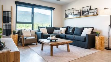 A modern living room with a dark grey sofa, a wooden coffee table, and a large window with a mountain view.
