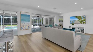 A spacious living area with a large sofa, a dining table with chairs, and a view of a swimming pool through glass doors. - Aqua Cottage- Newly renovated Park Shore home! (Naples)
