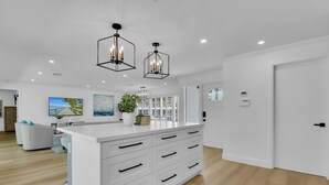 A modern kitchen with a central island, pendant lighting, and a view of the living area. - Aqua Cottage- Newly renovated Park Shore home! (Naples)