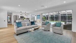 A modern living room with a sofa, coffee table, and armchairs, a kitchen in the background, and large windows offering a view of the outdoors. - Aqua Cottage- Newly renovated Park Shore home! (Naples)