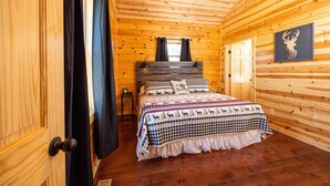 A wooden cabin bedroom with a bed, curtains, a window, and a deer head decoration. - Bourbon Trail Lodge by The Inn & Spa at Cedar Falls (Logan)