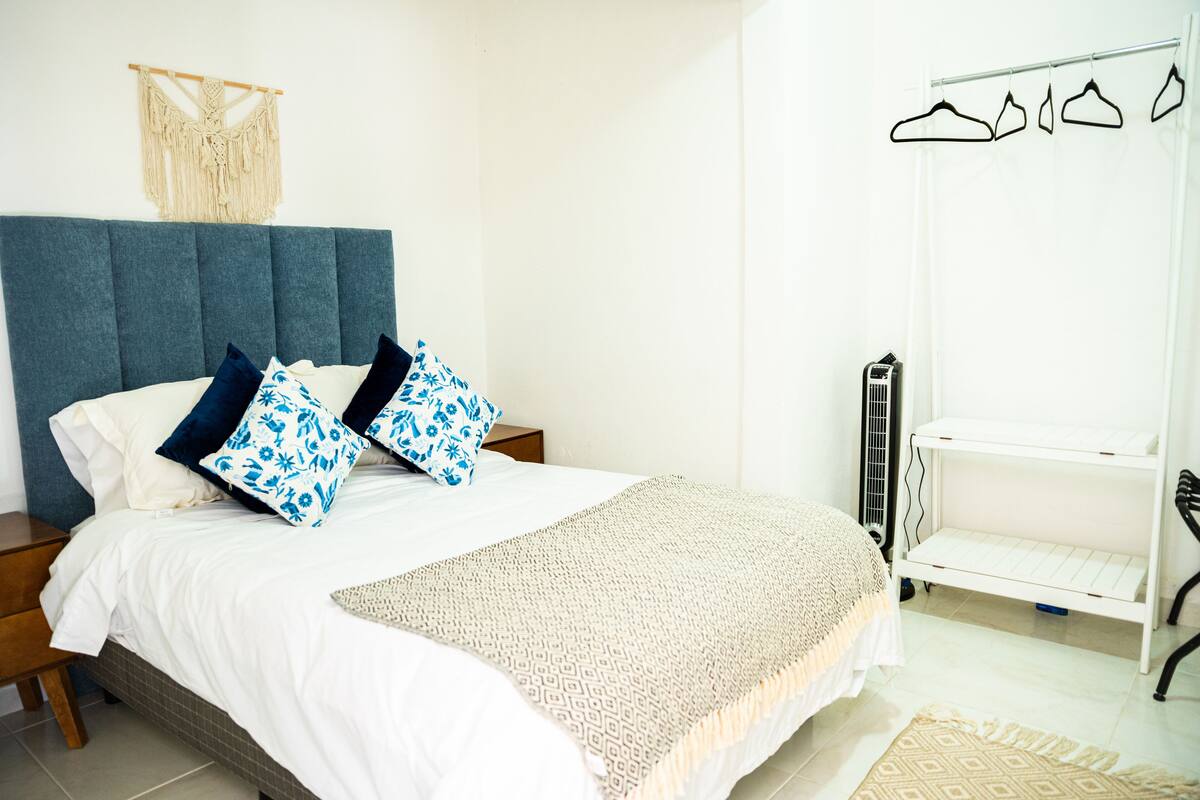 A neatly made bed with a blue headboard, white bedding, and patterned pillows. There is a white radiator and a white shelf unit to the right.