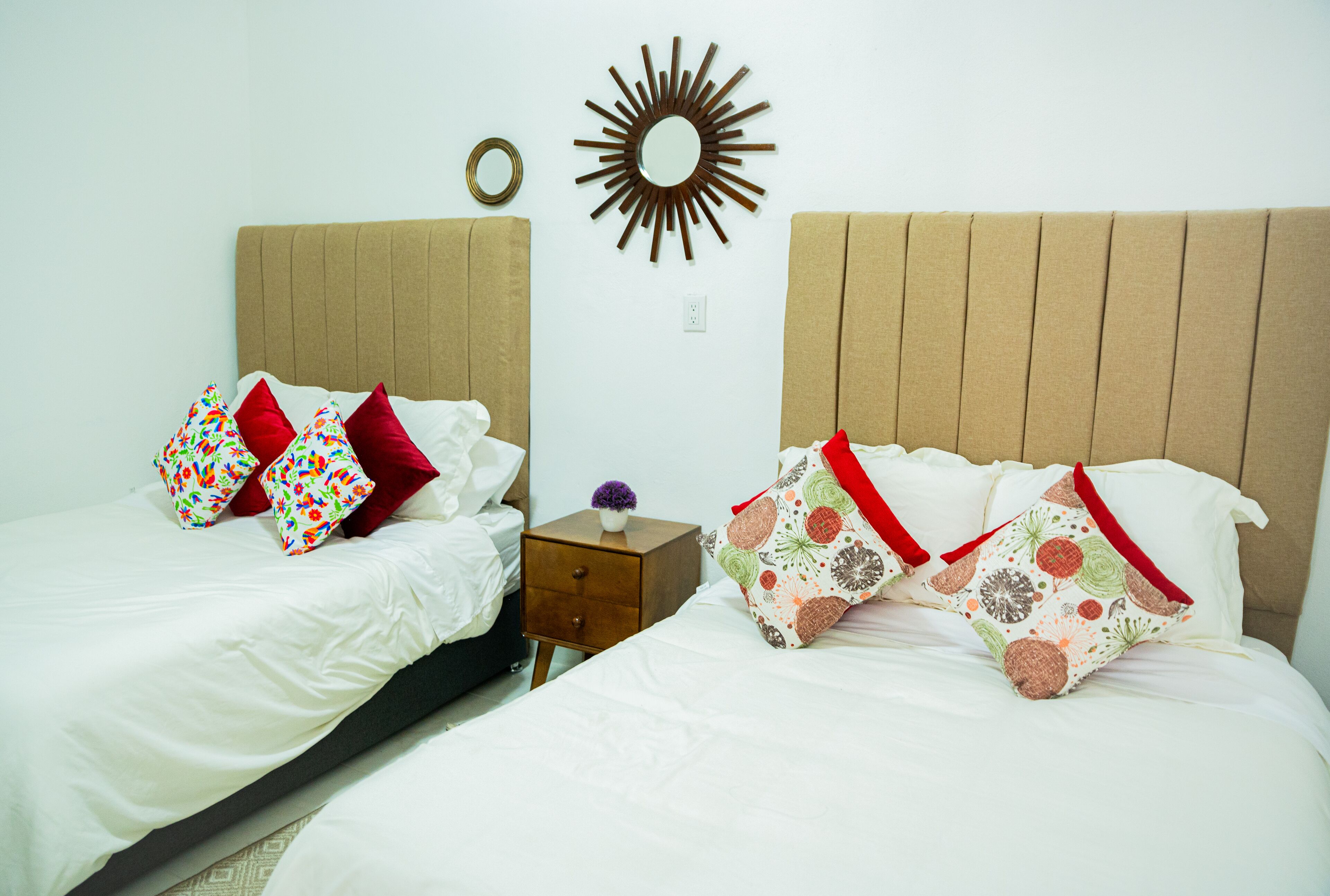 A double bed with floral-patterned pillows, a small bedside table with a vase, and a wall-mounted mirror.