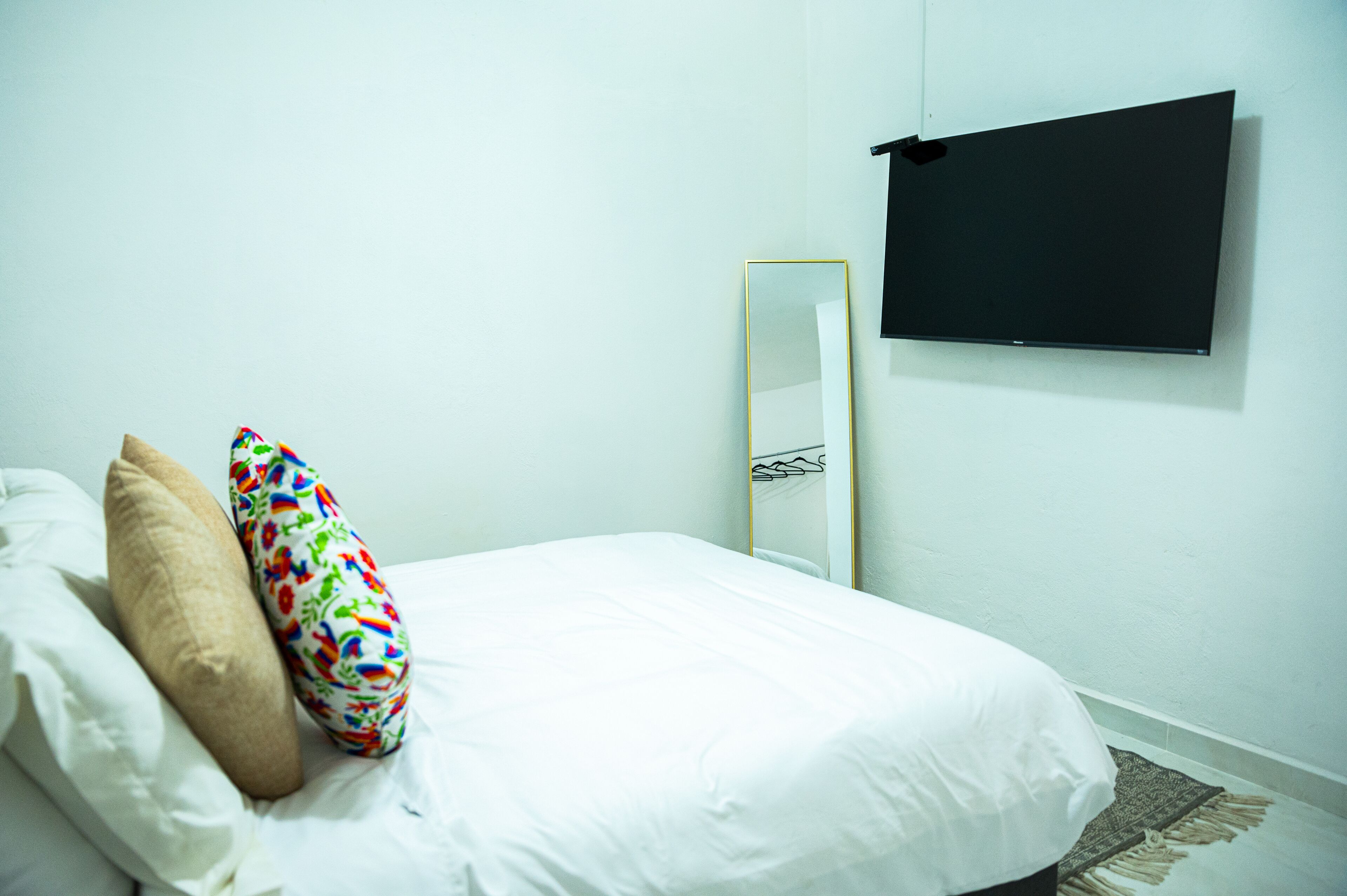 A bed with white bedding, two pillows, a colorful pillow, a black TV mounted on the wall, and a mirror.