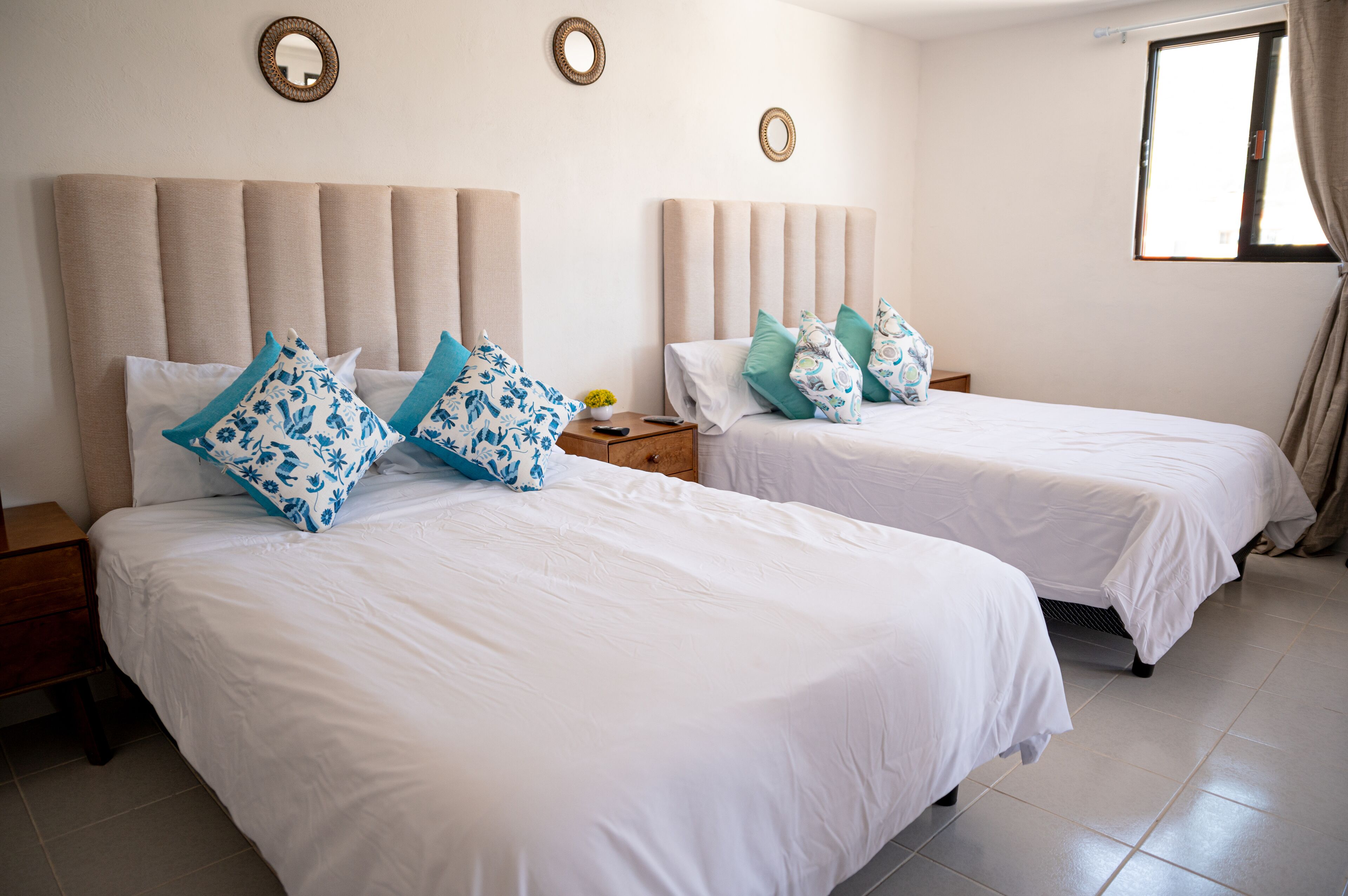 A hotel room with two beds, each with white bedding and blue patterned pillows. There are wooden nightstands with a small table and a lamp between the beds. The room has a large window with curtains, and the walls are adorned with circular wall art.