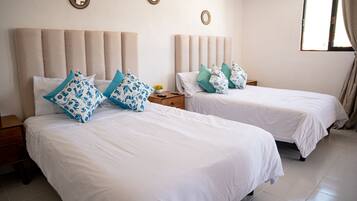 A hotel room with two beds, each with white bedding and blue patterned pillows. There are wooden nightstands with a small table and a lamp between the beds. The room has a large window with curtains, and the walls are adorned with circular wall art.