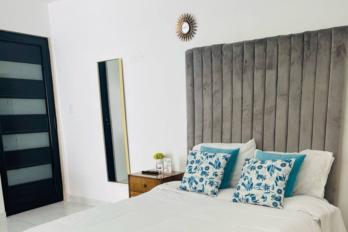 A modern bedroom with a large bed, a wooden headboard, a nightstand with a lamp, and a wall-mounted clock.