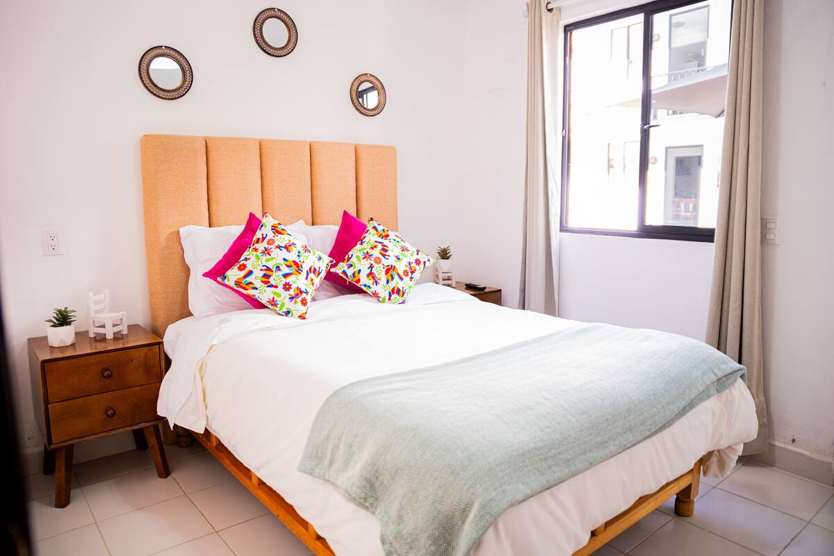 A neatly made bed with a light-colored headboard, two bedside tables with decorative mirrors, and a window with curtains.