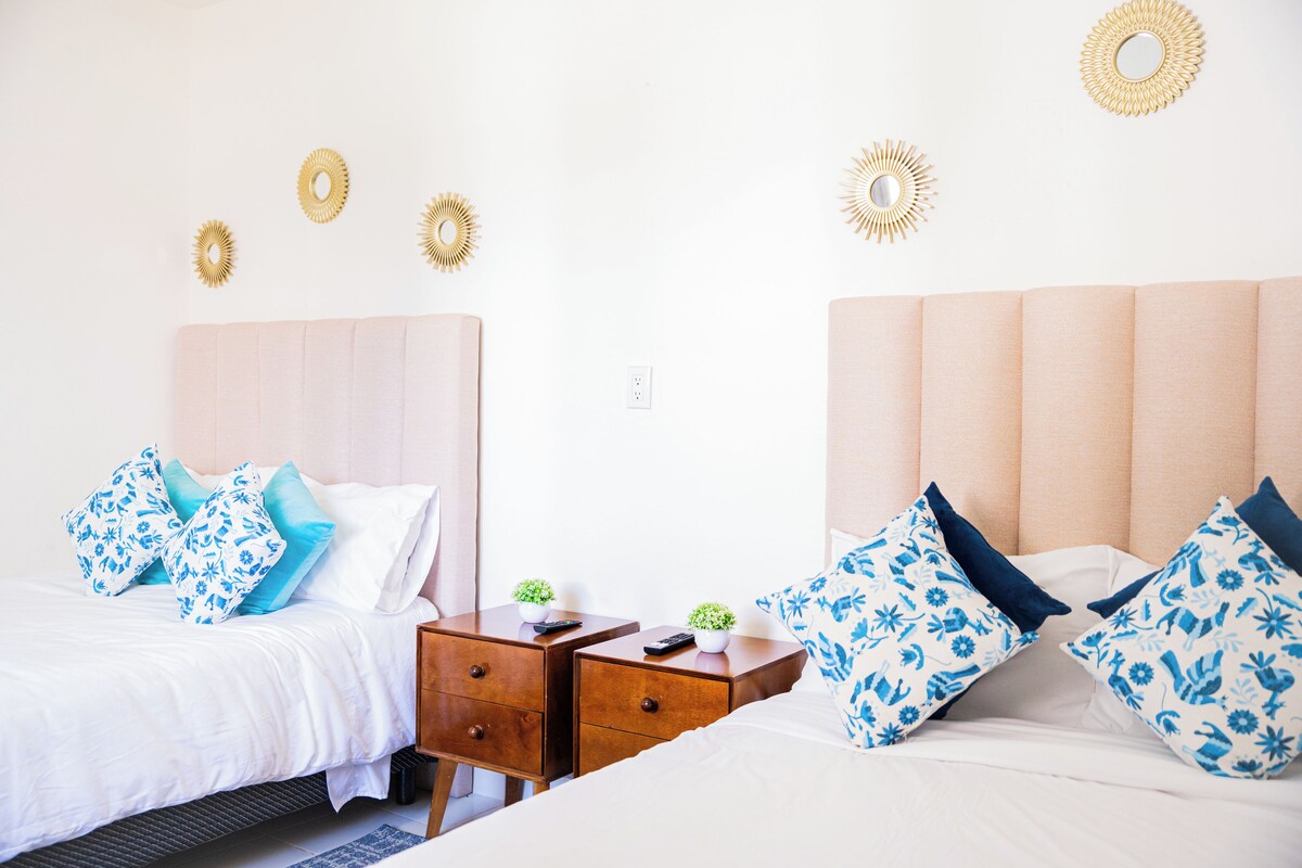 A hotel room with two beds, each with a headboard, a nightstand with a remote control, and decorative wall mirrors.