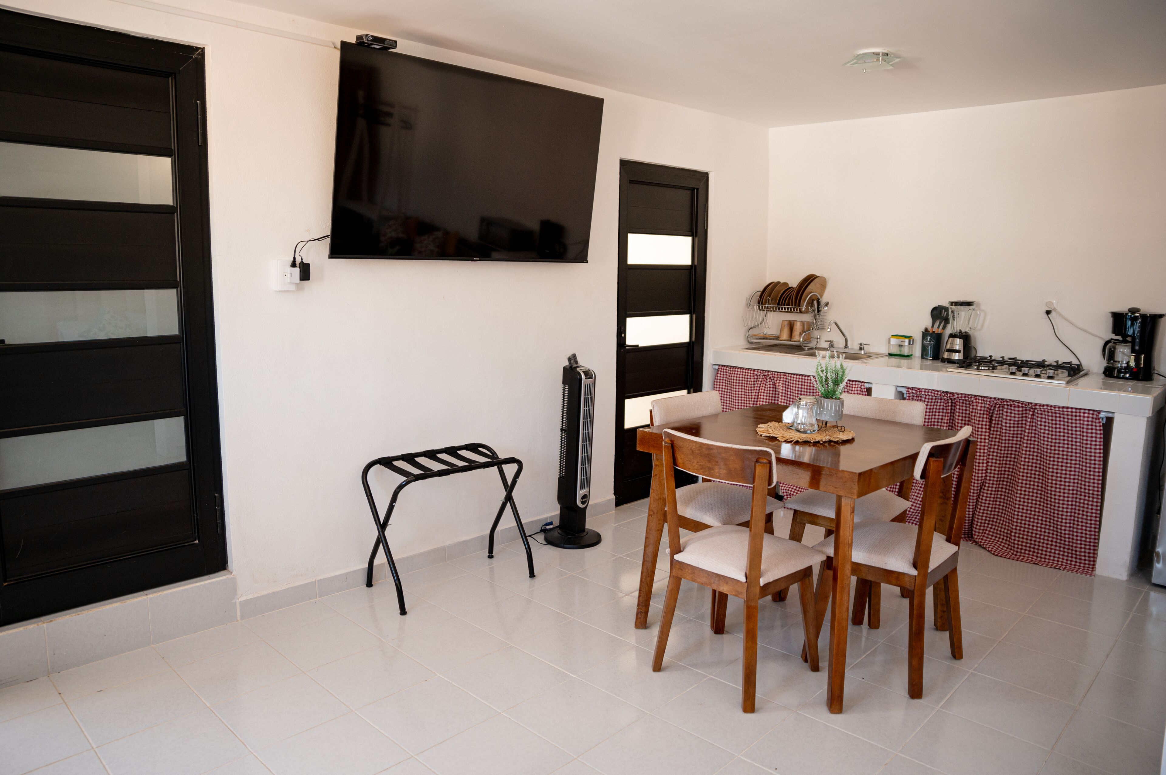 A compact kitchen with a dining area, a black-framed TV on the wall, and a small stool.