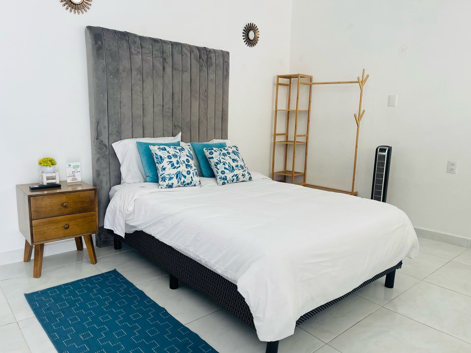 A modern bedroom with a large bed, a wooden nightstand, a small table with a plant, and a wall-mounted shelf.