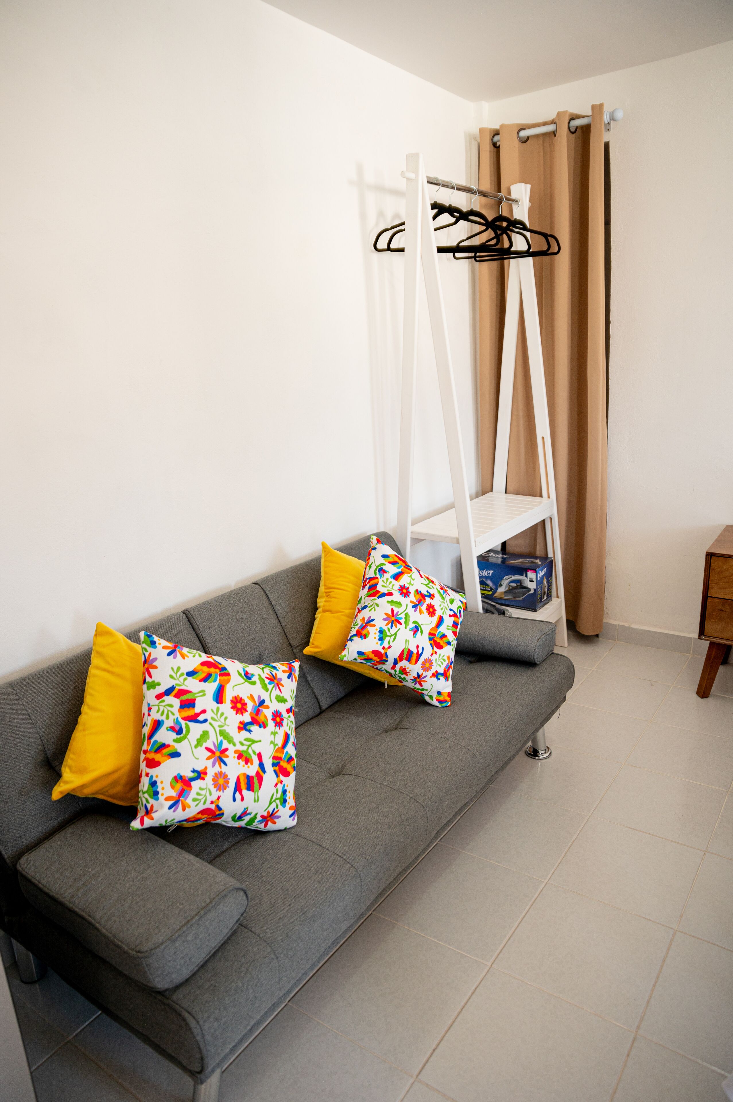 A grey sofa with yellow and floral patterned cushions, a white hanging rack, and a beige curtain.