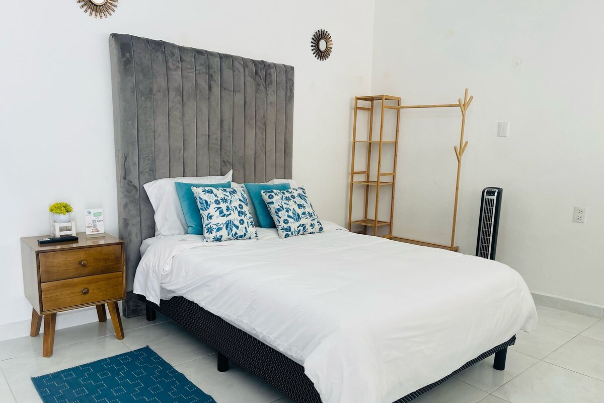 A modern bedroom with a large bed, a wooden nightstand, a small table with a plant, and a wall-mounted shelf.