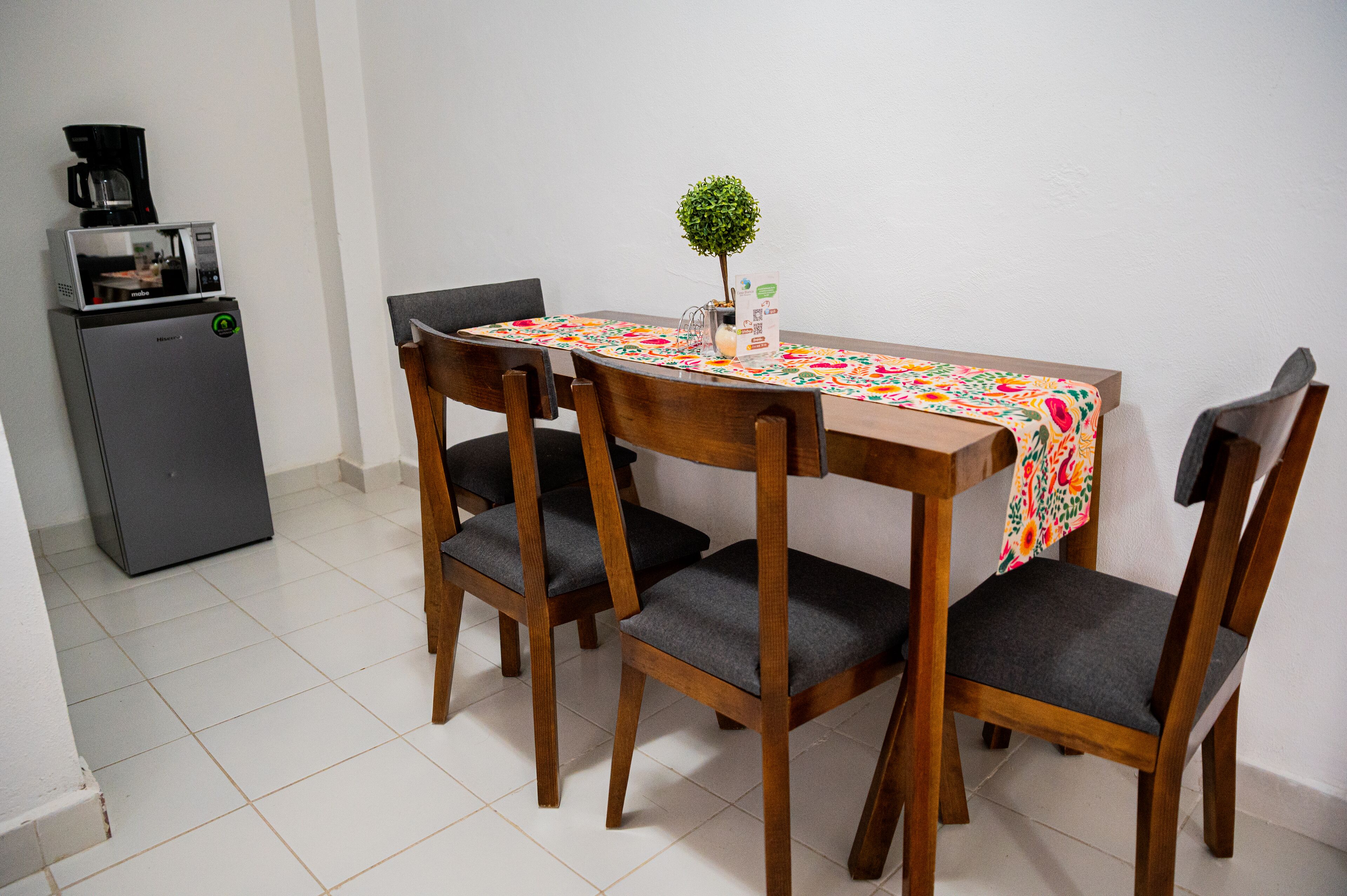 A dining area with a wooden table, four chairs, a small refrigerator, and a coffee maker.