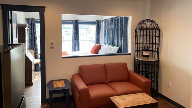 A compact living space with a brown sofa, a small wooden table, a window with curtains, a metal decorative piece, and a glimpse of a kitchen area. - Hyland Hotel (Palmer)