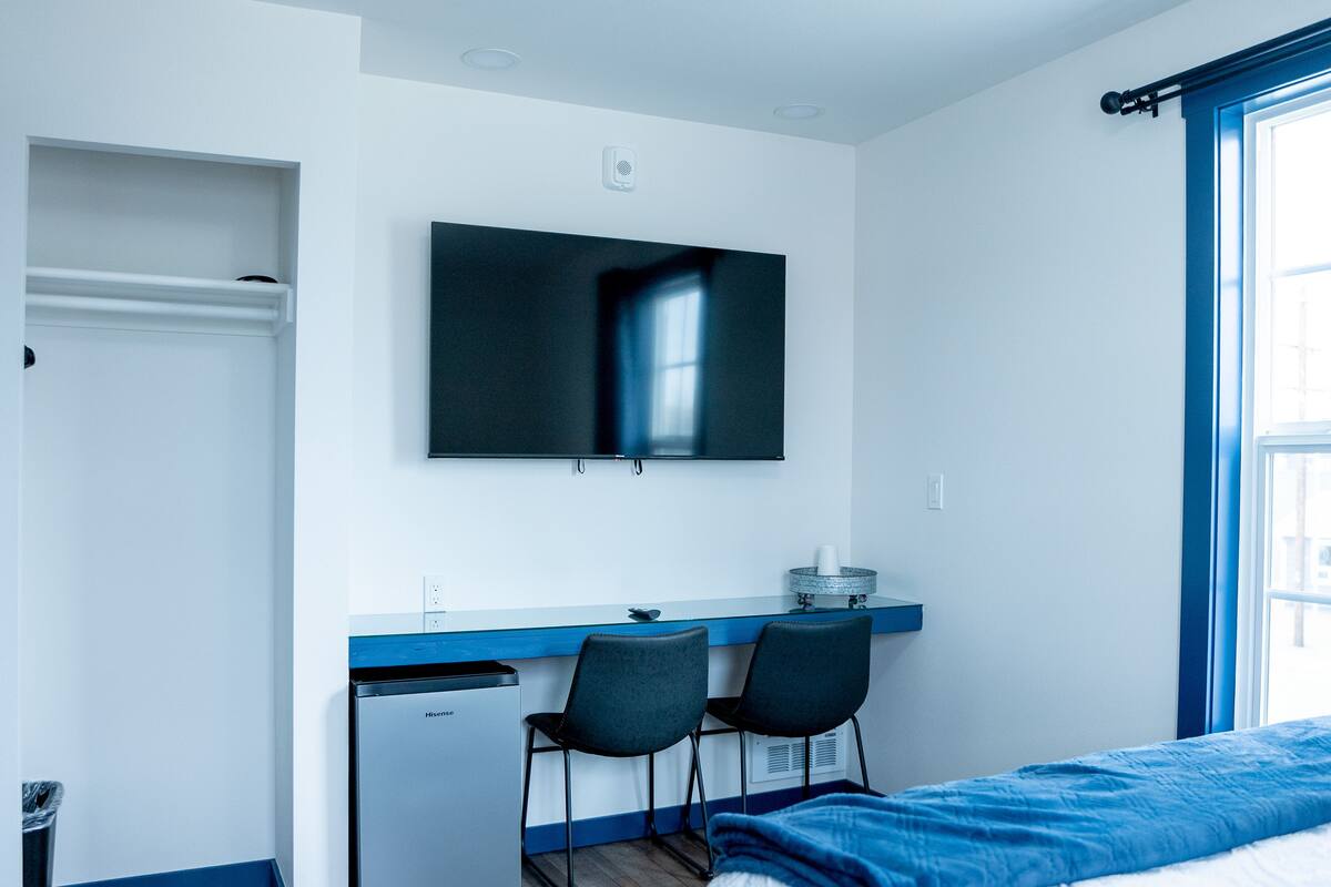 Moderni hotellihuone, jossa on sänky, työpöytä kahdella tuolilla, seinälle asennettu televisio ja ikkuna, jossa on verhot.