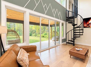 A spacious living room with a large window, a brown leather sofa, a wooden coffee table, and a black spiral staircase.