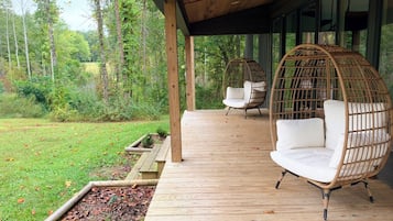 Eine Holzterrasse mit zwei Korbstühlen und einer gepolsterten Bank, mit Blick auf eine bewaldete Landschaft.