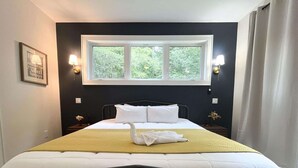 Una cama bien hecha con ropa blanca y una manta amarilla, una ventana con vista a la vegetación, y dos lámparas fijadas a la pared.