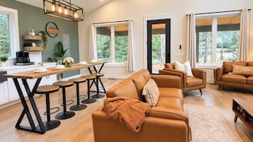 A modern living room with a large brown leather sofa, a wooden dining table with bar stools, and a kitchen area with a coffee maker.
