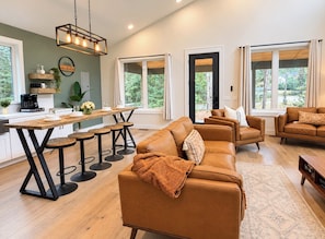 A modern living room with a large brown leather sofa, a wooden dining table with bar stools, and a kitchen area with a coffee maker.