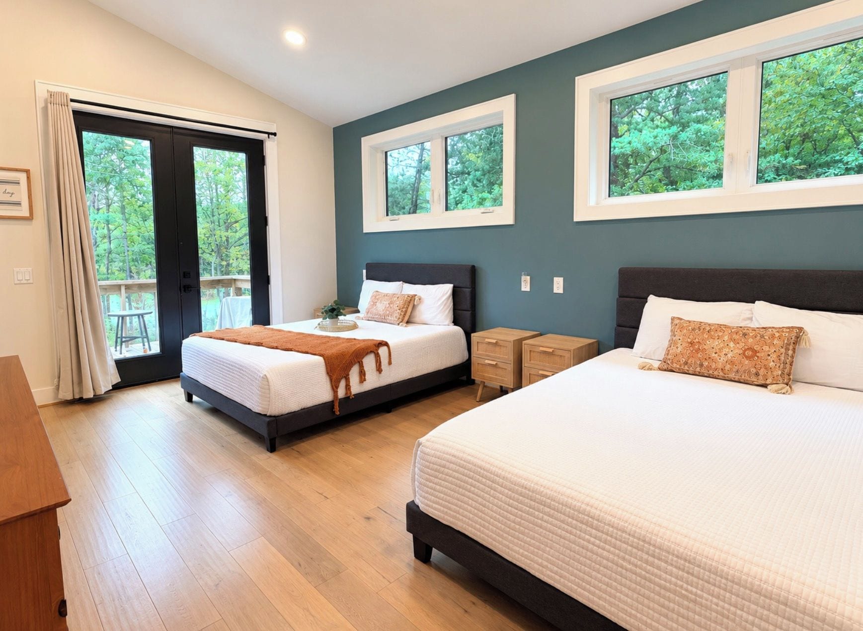 A bedroom with two beds, a wooden floor, a balcony door, and windows with a view of trees.