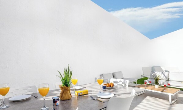 A sunlit outdoor dining area with white furniture, a table set with plates, glasses, and a plant.