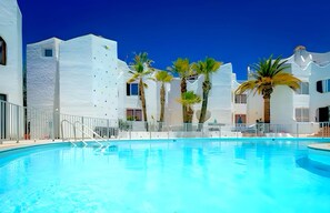 A community pool with a clear blue water surface, surrounded by white buildings and palm trees.