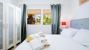 Ein Hotelzimmer mit einem Bett, einem Nachttisch mit roter Lampe, einem Fenster mit Vorhängen und Blick auf Palmen.