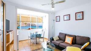 Ein Wohnzimmer mit einer Couch, einem Glastisch, einem Esstisch mit Stühlen, einem Fernseher und Meerblick durch große Fenster.