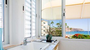 Ein Badezimmer mit einem weißen Waschbecken, einem Fenster mit Blick auf das Meer und Palmen sowie einer grünen Vase auf der Ablagefläche.