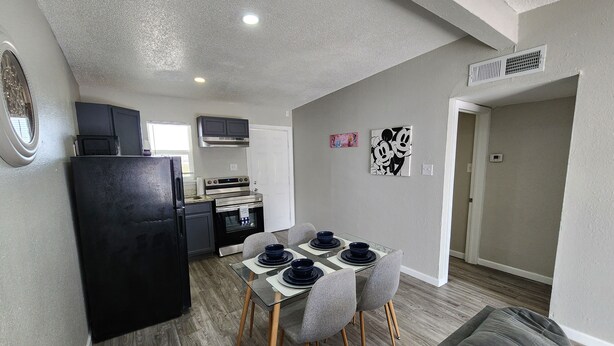 Una cocina funcional con electrodomésticos modernos, una zona de comedor con mesa de vidrio y sillas grises, y un sofá gris en un ambiente neutro.