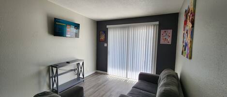 Una sala de estar con televisión de pantalla plana, un sofá, una mesa de centro y una ventana con persianas.