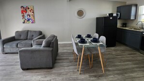 Una sala de estar moderna con un sofá gris, una mesa de comedor con platos azules, y una cocina con armarios oscuros y un refrigerador negro.