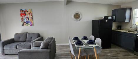 Una sala de estar moderna con un sofá gris, una mesa de comedor con platos azules, y una cocina con armarios oscuros y un refrigerador negro.