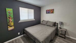 Un dormitorio con cama, mesita de noche, lámpara, ventana con persianas, un cartel en la pared y una lámpara en el techo.