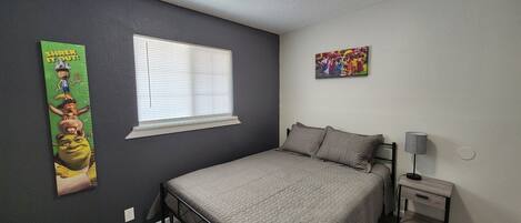 A bedroom with a bed, a nightstand, a lamp, a window with blinds, a poster on the wall, and a ceiling light.
